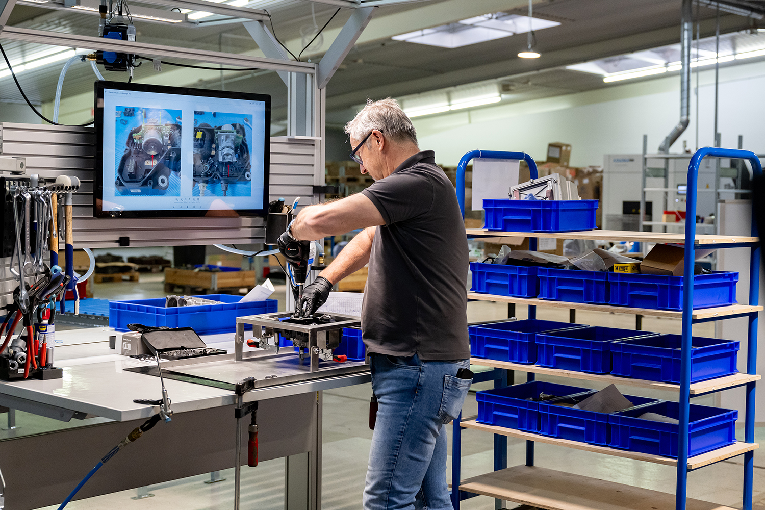 Ein Mann arbeitet an einem Montagearbeitsplatz in einer Fabrik und setzt dabei Werkzeuge ein, um Teile zusammenzubauen. Neben ihm stehen Regale mit blauen Aufbewahrungsbehältern, und über dem Arbeitsplatz zeigt ein Bildschirm technische Zeichnungen an.