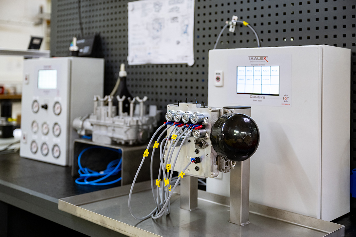 Ein Laborarbeitsplatz verfügt über moderne Bedienfelder, Messgeräte und Vorrichtungen, die mit Kabeln und Schläuchen verbunden sind und vor einer Lochwand stehen, an der technische Diagramme angeheftet sind.