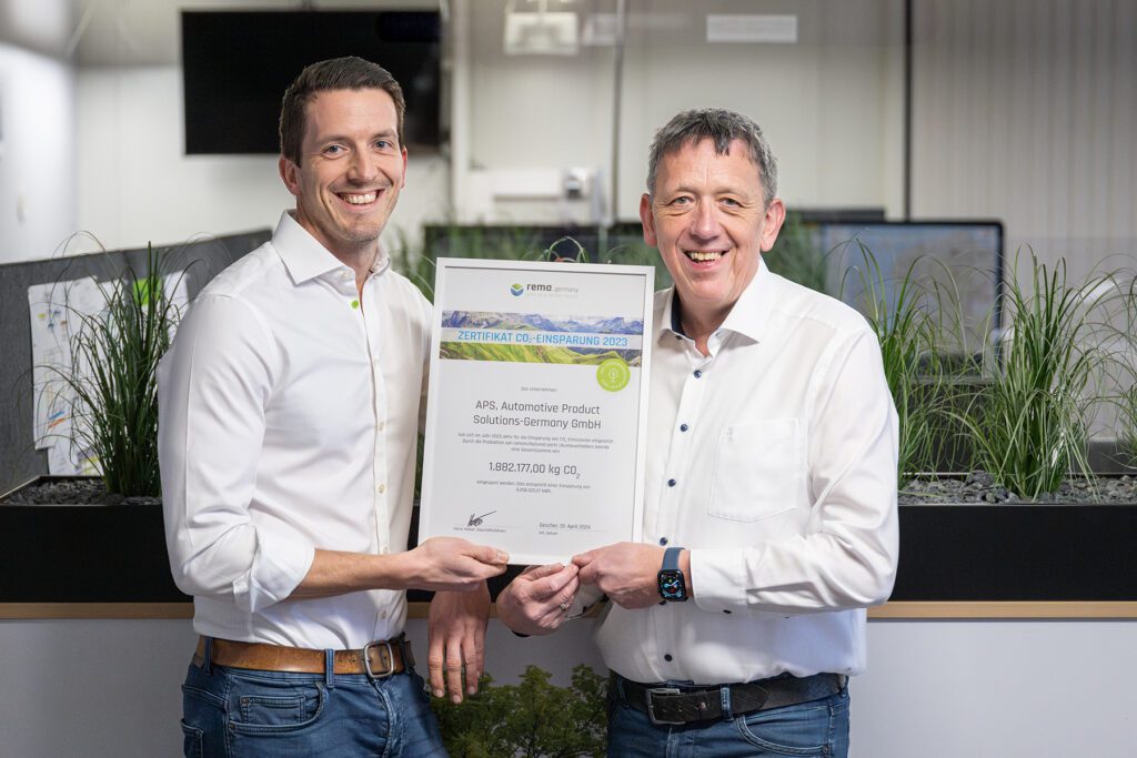 Twee mannen in witte overhemden glimlachen terwijl ze samen binnenshuis een certificaat vasthouden. Op de achtergrond zijn groene planten en computers te zien.