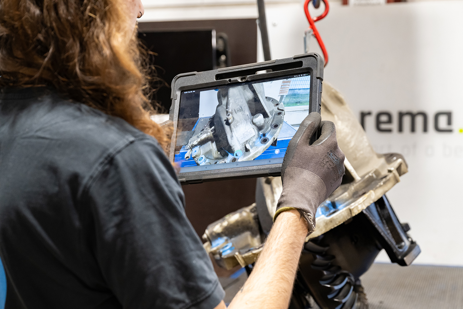 Eine Person mit langen Haaren und Handschuhen hält ein Tablet in der Hand und macht ein Foto oder ein Video von einem mechanischen Bauteil in einer industriellen Umgebung.