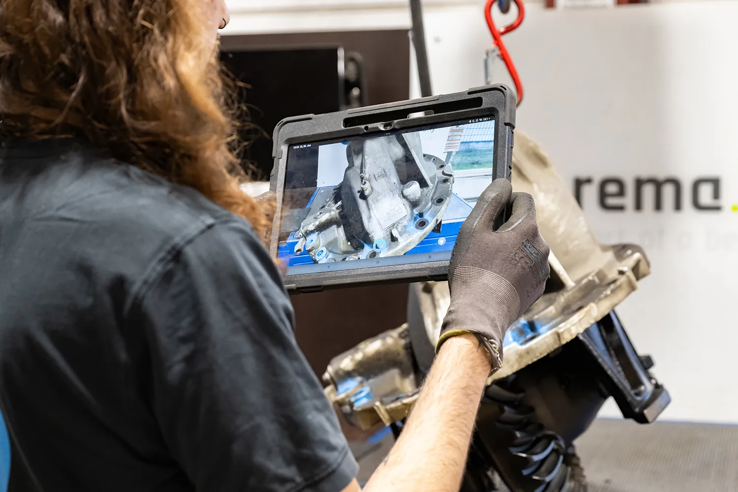 A person wearing gloves holds a tablet showing a mechanical component and examines it closely while standing next to the actual machine in a workshop.