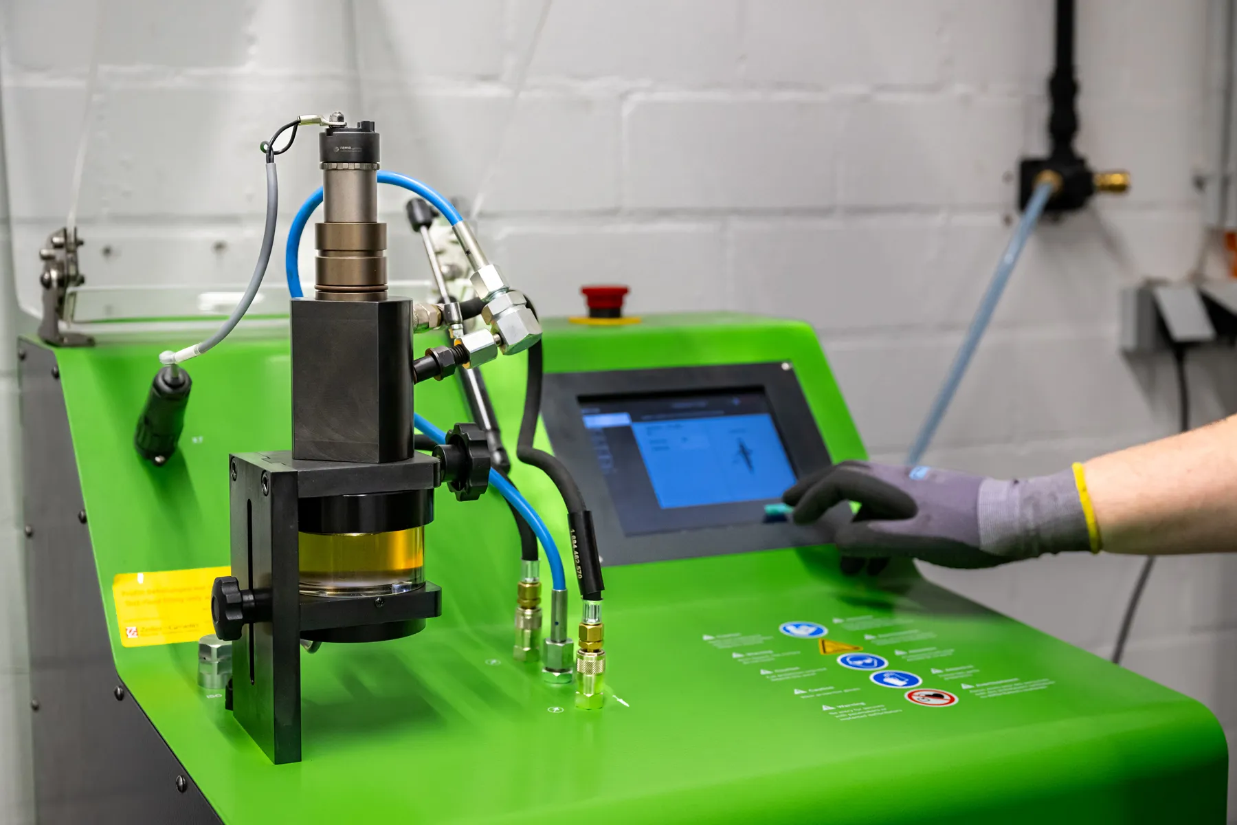 A person wearing a glove operates a green industrial machine with a touchscreen and several blue and yellow cables connected to the device.