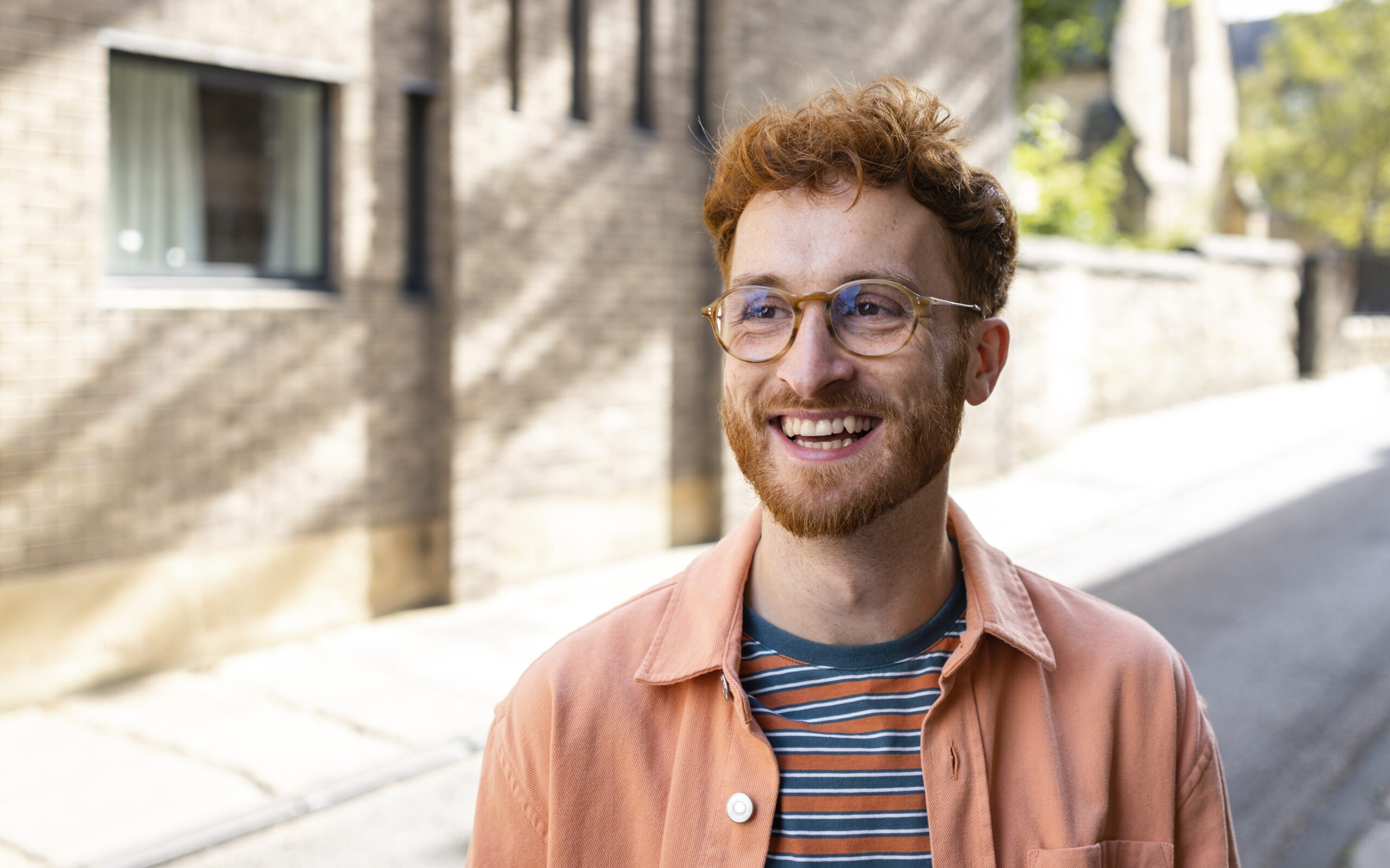 Eine lächelnde Person mit lockigem rotem Haar, Brille und Bart trägt eine pfirsichfarbene Jacke über einem gestreiften Hemd und steht an einem sonnigen Tag im Freien neben einem Backsteingebäude.